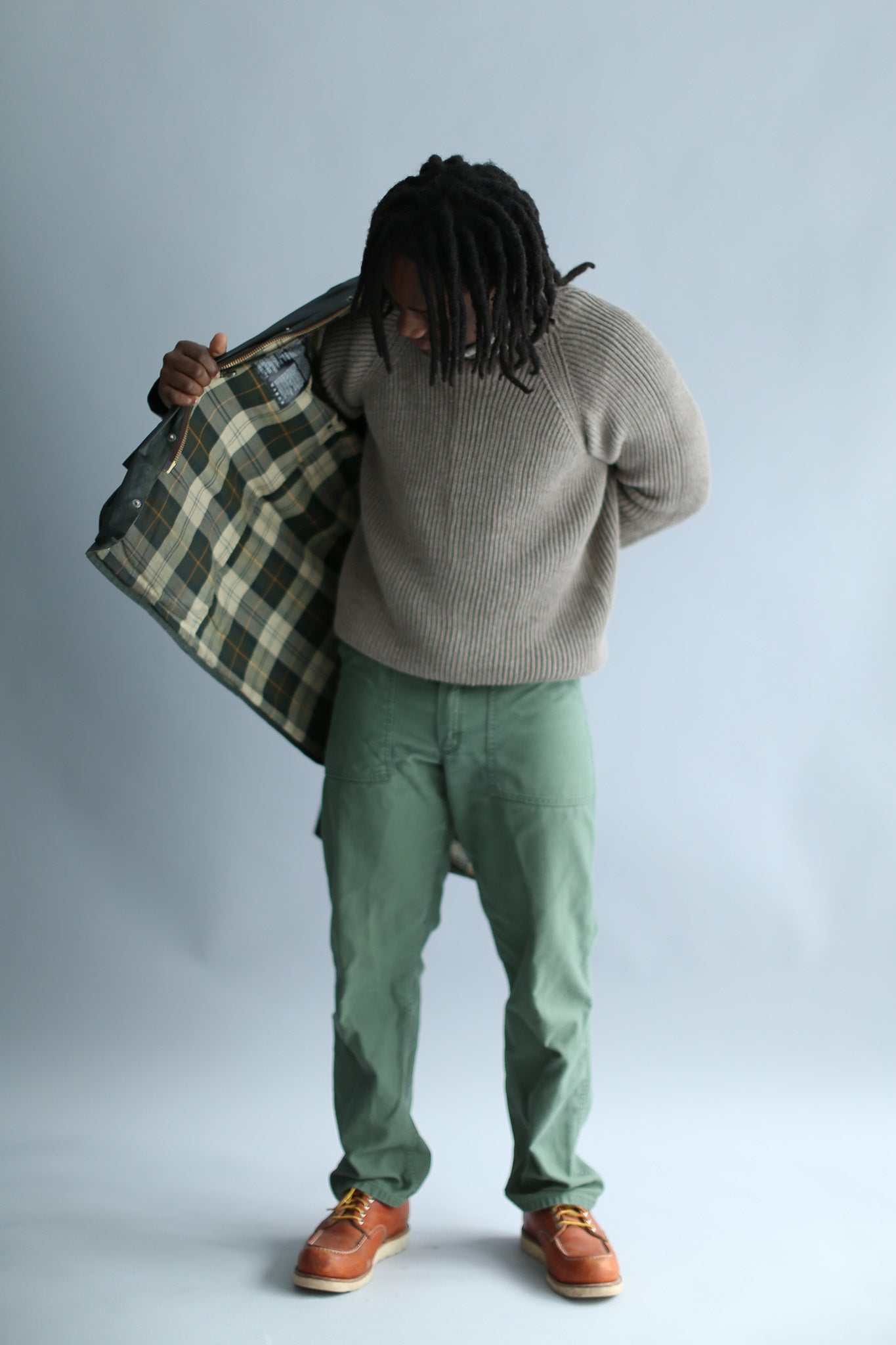 A man wearing a light brown ribbed turtleneck against a white background, putting on a waxed jacket