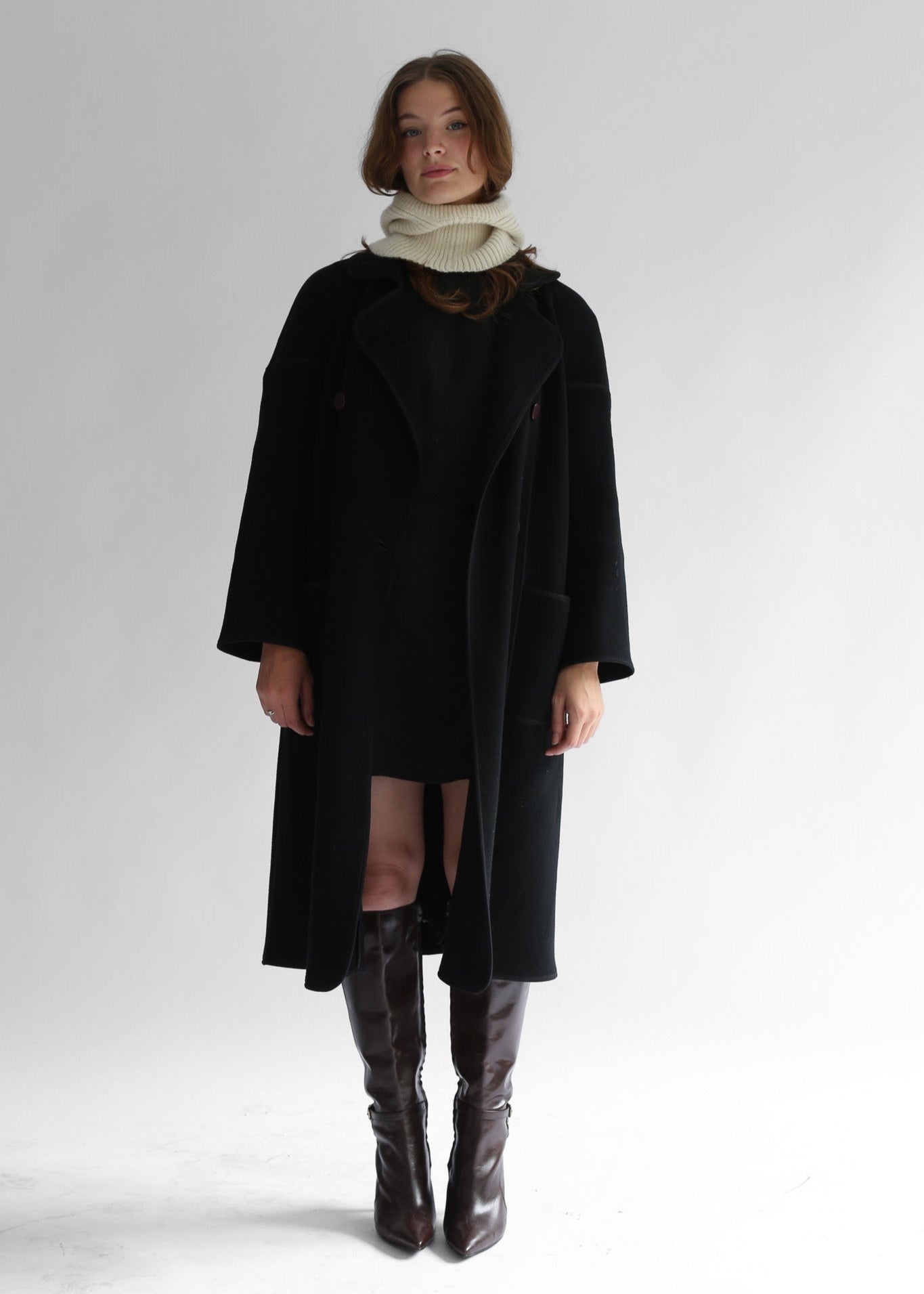 A model wearing a dark coat, skirt, boots and a white balaclava on a white background