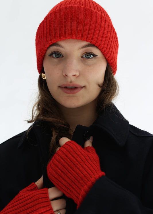 A woman wearing a bright red ribbed knit beanie, matching fingerless gloves, and a black coat, photographed against a clean white studio background.
