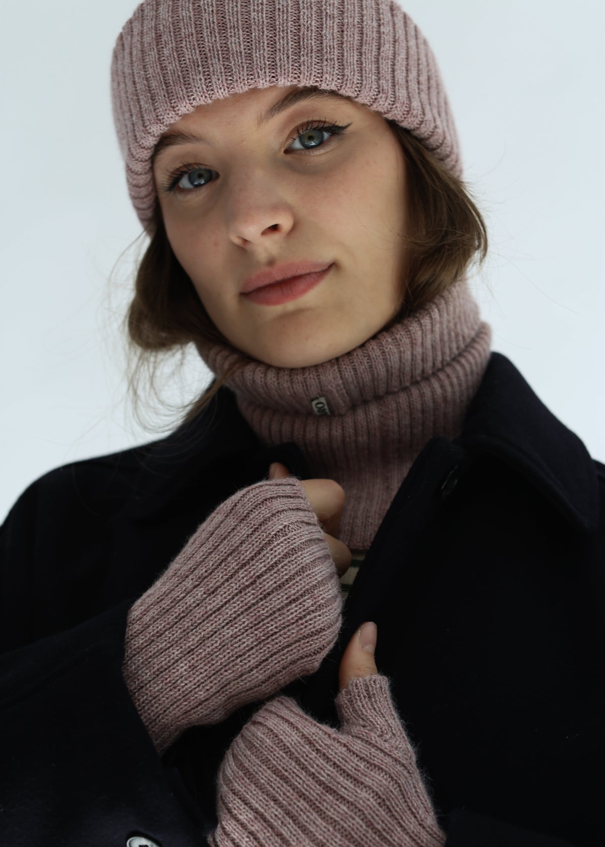 A close-up of a woman wearing a matching ribbed knit winter set consisting of a dusty rose beanie, turtleneck scarf, and fingerless gloves, paired with a dark coat against a light gray background.