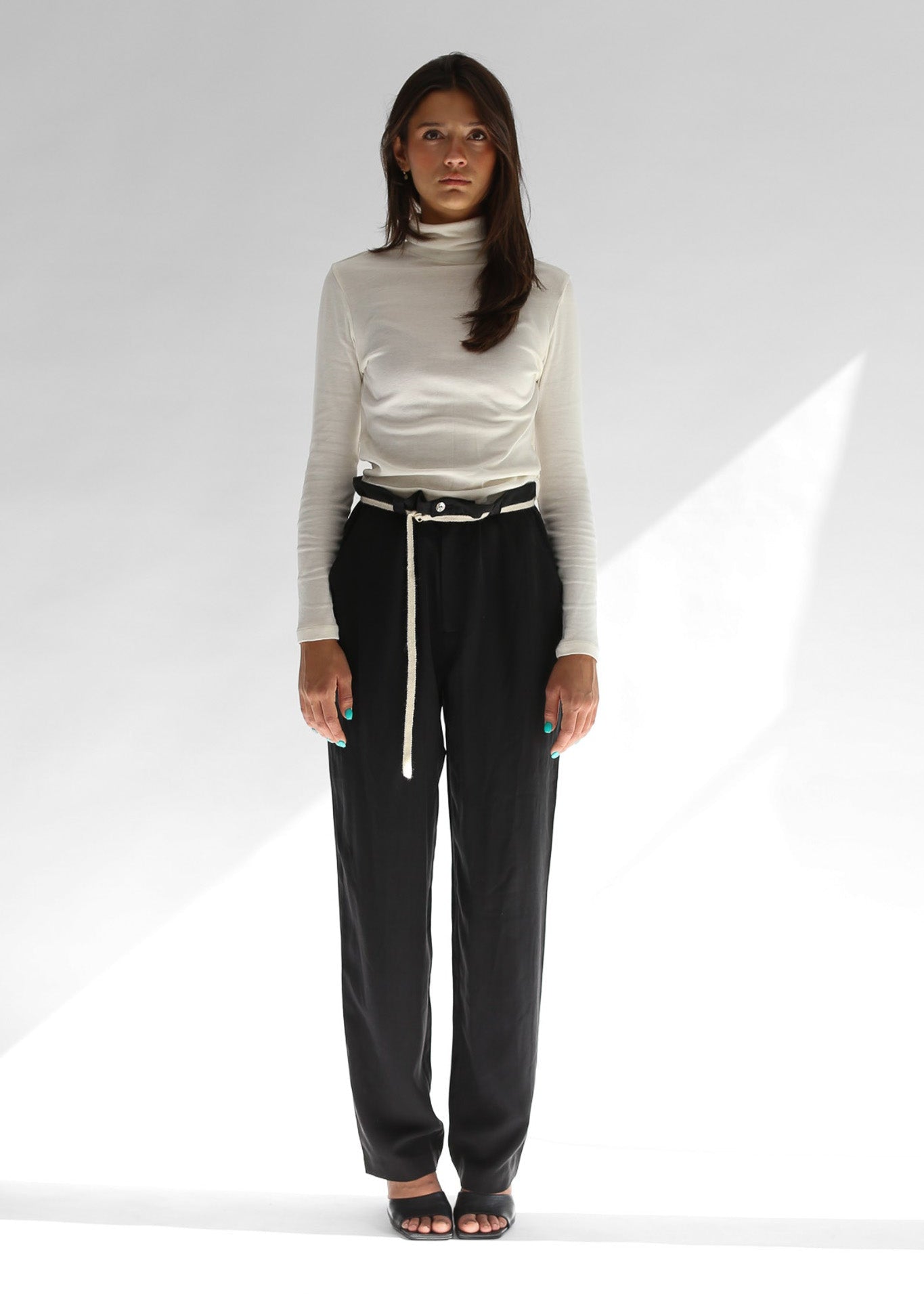 Woman wearing a white turtleneck and black pants with a white knit belt on a white background