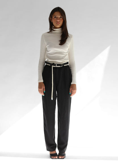 Woman wearing a white turtleneck and black pants with a white knit belt on a white background