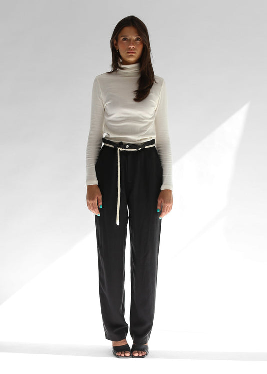 Woman wearing a white turtleneck and black pants with a white knit belt on a white background