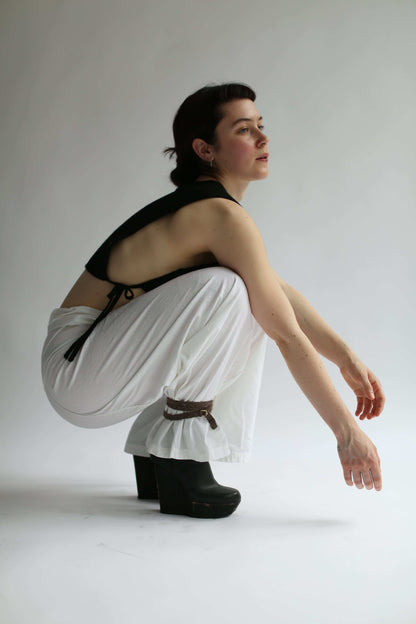 Woman squatting wearing a black knit top, white pants and a black ankle knit belt