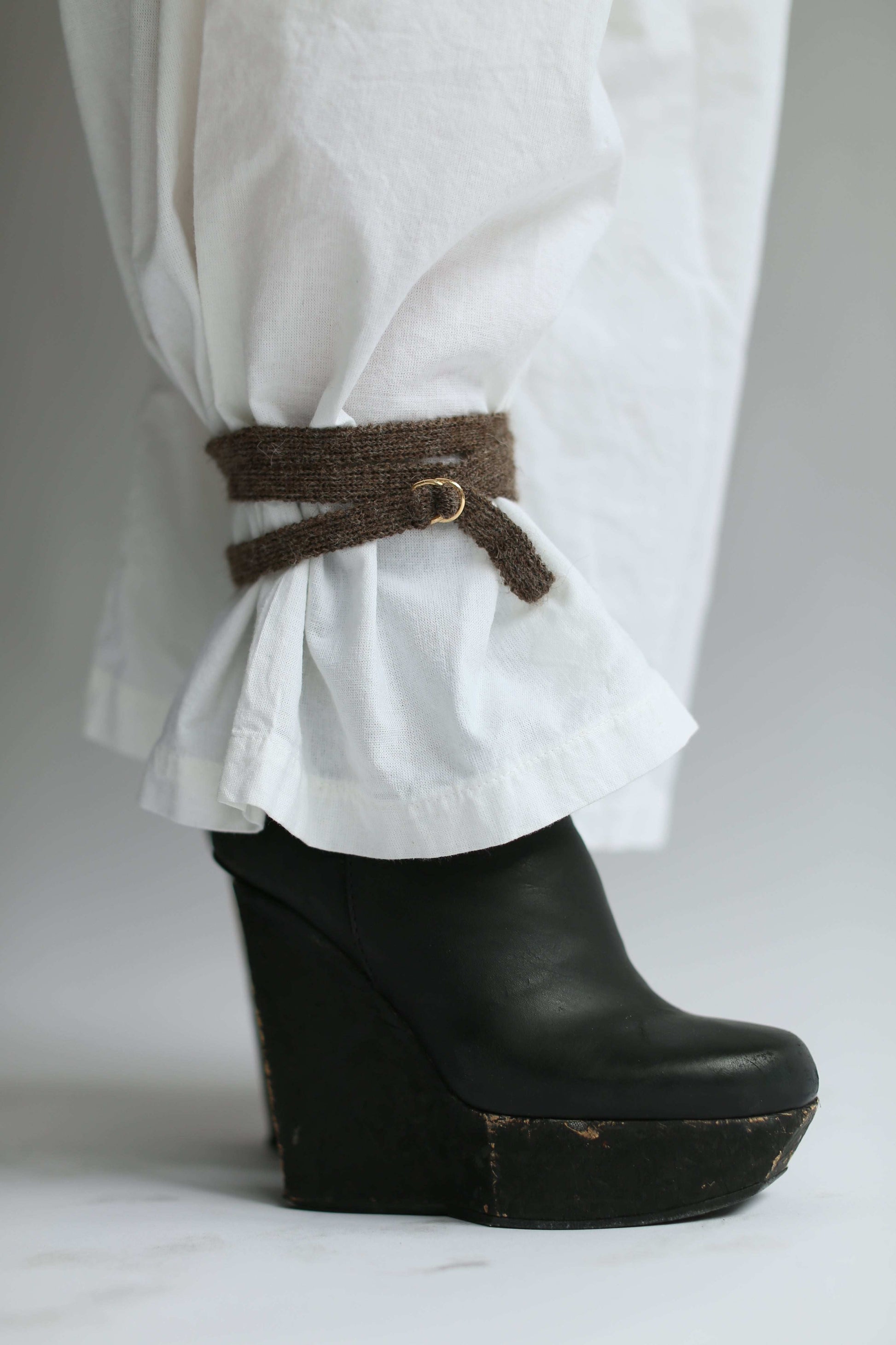 Detail of a brown knit belt at the ankle over white pants and black leather shoes